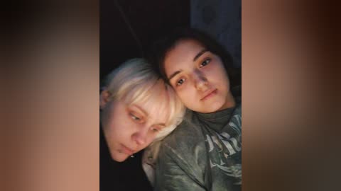 Media: A video of two young women with light skin, one with platinum blonde hair, the other with dark hair, both wearing casual clothing, lying close together on a dimly lit bed with a patterned headboard.