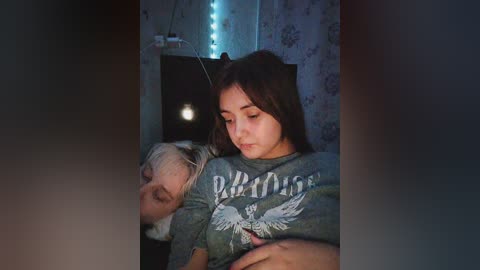 Media: Video of a young girl with long brown hair, wearing a gray \"Pheonix\" t-shirt, lying on a bed next to an elderly woman with short gray hair. The room features blue LED lights and floral wallpaper.