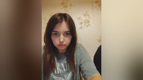 Media: Video of a young woman with long brown hair, wearing a grey t-shirt with a graphic, in a room with floral wallpaper.