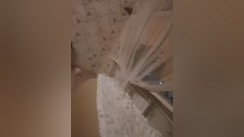 Video of a close-up wedding cake with a tiered, white fondant base and delicate, flowing white fabric draped over the top, surrounded by a beige wall. The image is slightly blurry.