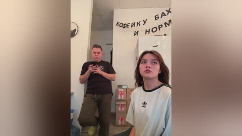 Media: Video of a man in casual clothes taking a selfie, with a woman in a white Adidas shirt and brown hair in the background.