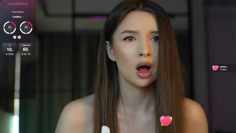 Media: Video of a young, fair-skinned woman with long, straight brown hair, wearing a white top, in a blurred modern room. She has an open-mouthed expression, with heart emojis and virtual meters in the corner.