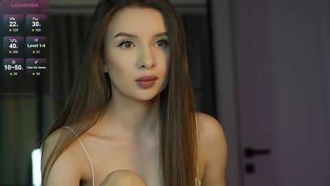 Media: A video of a young woman with long brown hair, wearing a light-colored spaghetti strap top, sitting in a dimly lit room.