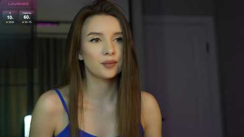 Media: Video of a young Caucasian woman with long brown hair, wearing a blue sleeveless top, standing indoors, with a blurred background.