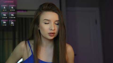 Media: A video of a young woman with long brown hair, fair skin, and closed eyes, wearing a blue sleeveless top, indoors with dim lighting.