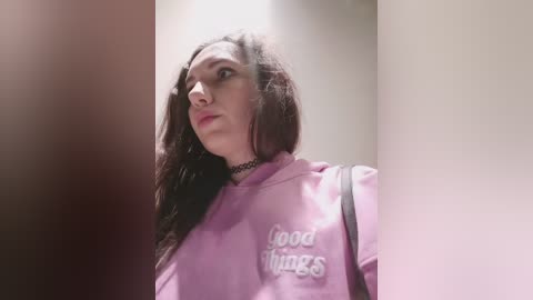 Video of a young woman with long, dark hair, wearing a pink hoodie with \"good vibes\" text, looking contemplative, with a black choker. Background is blurred.