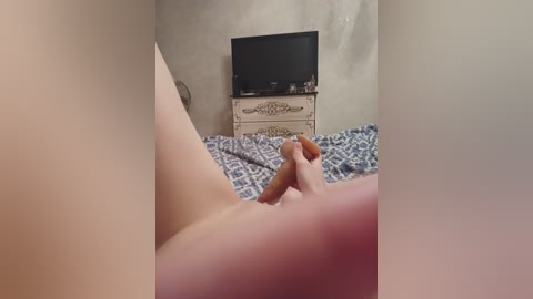 Video of a nude woman lying on a bed, holding her erect penis, in a dimly lit bedroom with a beige wall, a patterned bedspread, and a small TV on a dresser.