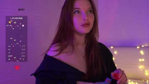 A video of a young woman with long brown hair, wearing a black off-shoulder top, standing indoors with a purple background.