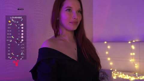 Video of a young Caucasian woman with long, straight red hair, wearing a black off-shoulder top, standing in a softly lit bedroom with purple and yellow fairy lights, a \"LOVE\" wall sign, and a digital clock.
