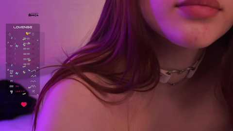 A close-up video of a young woman with long, wavy brown hair and fair skin. She wears a white choker necklace. The background is dimly lit with purple and pink hues, showcasing a \"Love Live!\" smartphone app interface on the left.