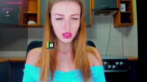Video of a fair-skinned, blonde woman with long hair, wearing a turquoise off-shoulder top, blowing a kiss in a modern kitchen with wooden cabinets, white tiles, and a black stove.