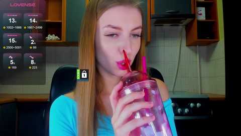 Video of a blonde woman in a turquoise top drinking from a pink straw in a modern kitchen.