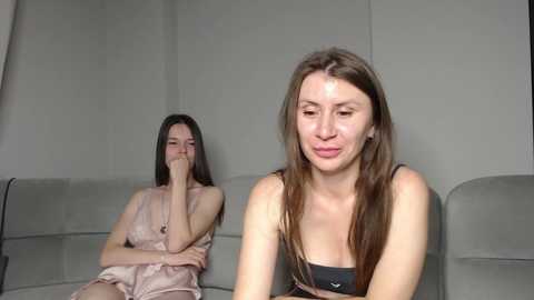 Video of two young women in a living room. One sits with a distressed expression, wearing a pink dress. The other, in a black top, looks annoyed.