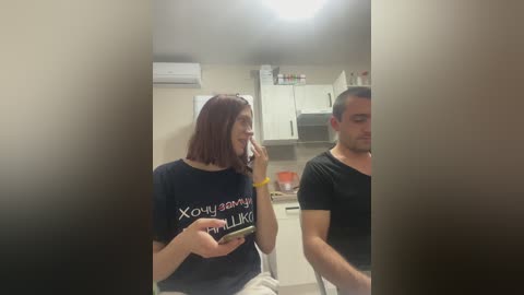 Video of a woman with shoulder-length auburn hair, wearing a black t-shirt with \"XOXO BOYFRIEND\" in white text, talking on a phone in a modern kitchen.