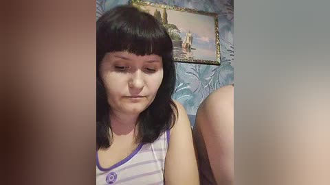 Video of a young woman with straight black hair, wearing a purple-striped tank top, sitting in a room with a framed painting of a beach scene on the wall.