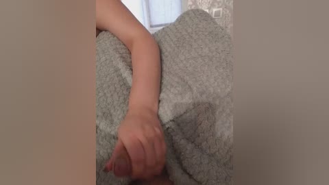 Video of a person's hand clutching a hard, erect penis under a light grey, fuzzy blanket. The background is a beige wall with a window and a power outlet. The setting appears intimate and private.