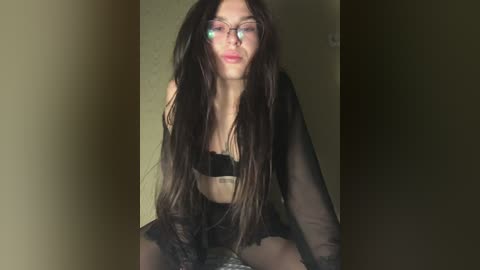 Video of a young woman with long, dark hair, wearing black lingerie, glasses, and sheer black robe, posing indoors.