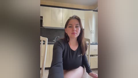 Media: Video of a young woman with light skin and straight brown hair, sitting in a modern kitchen with white cabinets and a black countertop. She wears a black long-sleeve shirt and beige shorts.