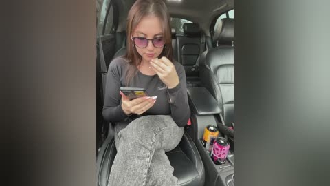 Media: Video of a young woman with glasses, seated in a car, eating a snack while texting, wearing a black top and grey pants.