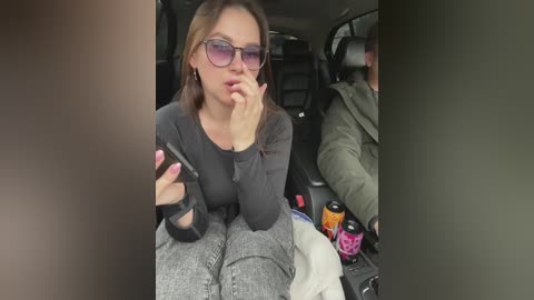 Media: Video of a woman with light skin and shoulder-length brown hair, wearing glasses, a black top, and grey pants, sitting in a car. She has a phone to her ear and a drink next to her.