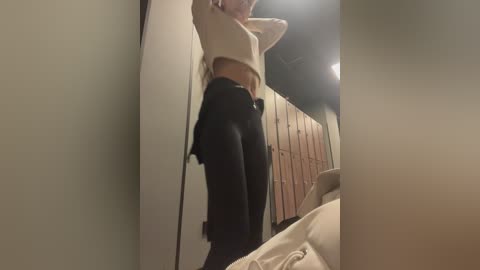 Media: A video of a slim, fair-skinned woman in a beige sweater and black leggings, standing in a dimly lit room with a wooden wardrobe and bed visible.