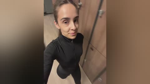 Media: Video of a young woman with light brown skin, straight brown hair, and a slender build, wearing a black zip-up jacket, standing in a locker room with wooden lockers.