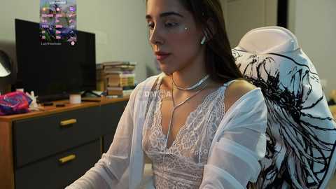 Media: Video of a young woman with light brown skin, dark hair, and light makeup, wearing a white lace bralette and sheer cardigan, seated in a gaming chair, with a TV and desk in the background.