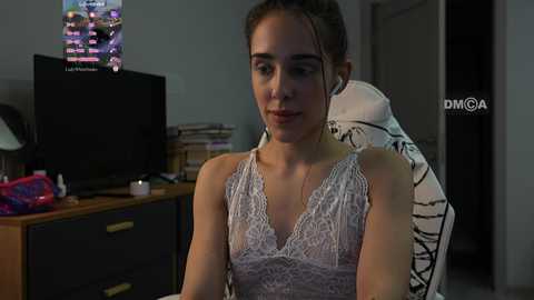 Media: Video of a young woman with light skin and brown hair, wearing a white lace camisole, seated at a desk in a dimly lit room with a computer screen and bookshelf background.