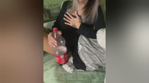 Video of a woman with light skin, long blonde hair, wearing a black long-sleeved top and grey patterned pants, sitting on a green quilted bedspread. She holds a red Coke bottle, one hand on her chest. Background includes a green wall and a white towel.