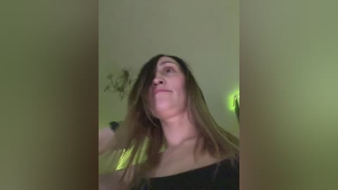 A video of a woman with long, brown hair, wearing a black off-shoulder top, standing in a dimly lit room with green walls.
