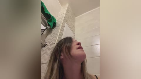 A video of a young woman with long, straight, light brown hair, looking upwards in a tiled bathroom. Green towel and white toiletries hang on the wall.