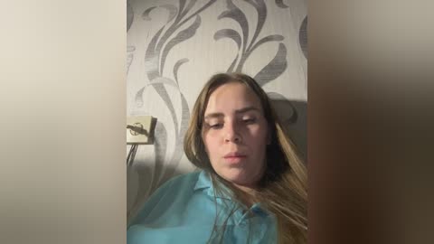 Media: Video of a Caucasian woman with long brown hair, wearing a light blue blouse, looking down in a dimly lit room with a gray and white patterned wallpaper.