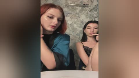 Media: Video of two women with fair skin and long hair. One with red hair in a teal blouse, the other with black hair in a black lace top, both looking contemplative in a bathroom with floral wallpaper.
