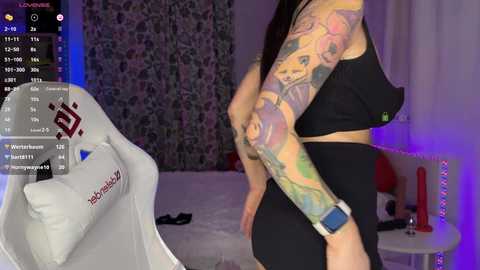 Media: Video of a tattooed woman in black sports bra and shorts, standing in a dimly-lit bedroom with a floral curtain, purple lighting, and a white gaming chair in the background.