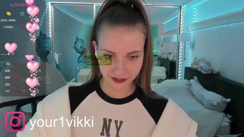 Media: A video of a young woman with light skin and long brown hair, wearing a white and black NY Yankees baseball shirt, in a brightly lit bedroom with hearts overlaying the image.