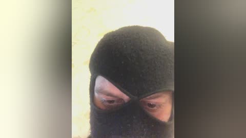 A close-up video of a person with a dark hood covering their face, revealing only their eyes. The background is a blurred, off-white wall. The image conveys a sense of mystery and secrecy.