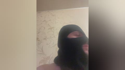A video of a person wearing a black balaclava, standing in front of a beige wall with a floral pattern. The image is slightly blurry, emphasizing a sense of secrecy.