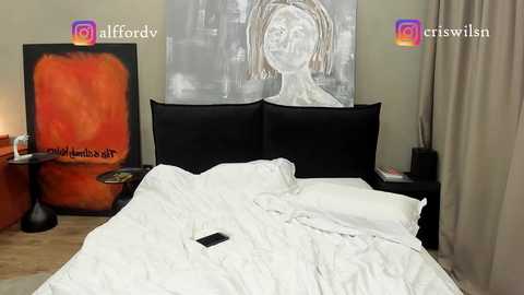 Media: Video of a modern bedroom with a black upholstered headboard, a white duvet, and two pillows. A large, abstract painting in shades of orange and black hangs behind the bed.