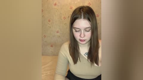 Media: Video of a young Caucasian woman with straight brown hair, fair skin, and light makeup, wearing a beige top and black skirt, seated indoors with a floral wallpaper backdrop.