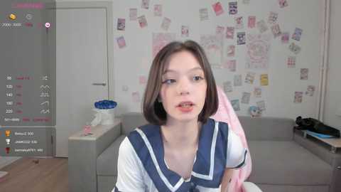 Video of a young woman with short brown hair, wearing a blue sailor school uniform, sitting in a living room with a cluttered, pink wallpapered wall.