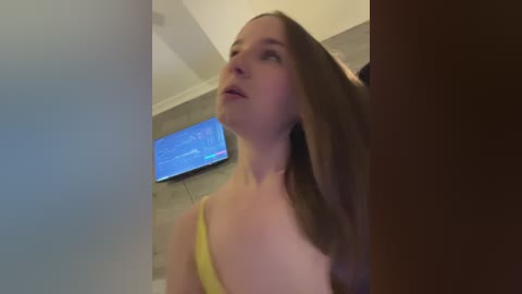 Video of a young woman with long, straight brown hair, wearing a yellow tank top, looking upward, standing in a modern, well-lit room with a flat-screen TV on the wall.