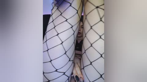 Media: Video of a woman's legs and face partially obscured by black fishnet stockings, showing a peek of her lips and nails.