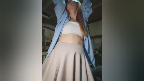 Video of a young woman with fair skin and long blonde hair, wearing a white bandeau top and light gray pleated skirt, posing with arms raised in a blue shirt. Background shows a cluttered room with shelves and boxes.