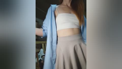 Video of a young woman with long brown hair, wearing a white crop top, beige skirt, and open blue shirt, standing in a dimly lit room with cluttered background.
