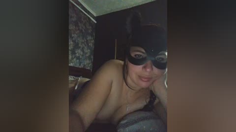 Media: Video of a topless woman with light skin, wearing a black cat mask and lingerie, lying on a bed with floral-patterned wallpaper in the background.