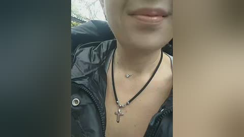 Video of a young woman with light skin, wearing a black jacket, black necklace, and a cross necklace, smiling slightly. Background is blurred.