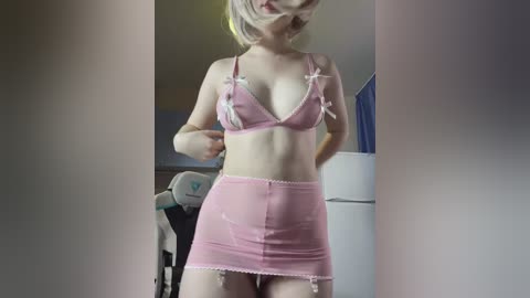 Video of a slim, blonde woman with pale skin, wearing a pink, sheer lingerie set with white lace trim and bows, standing in a dimly lit room with a white refrigerator and gaming chair in the background.