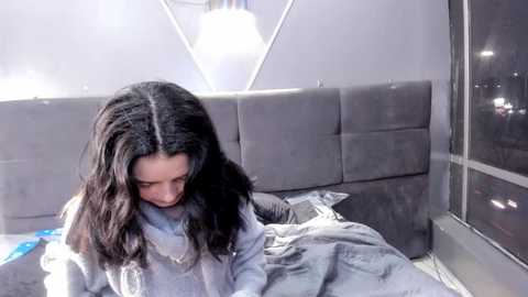 Video of a woman with long black hair, wearing a grey sweater, sitting on a bed with grey upholstered headboard, looking down, surrounded by a minimalist, modern room with a large window and a white, triangular pendant light.