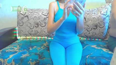 Media: Video of a woman in a bright blue sports bra and matching leggings, sitting on a teal couch with ornate, patterned pillows. She's holding a phone, smiling.