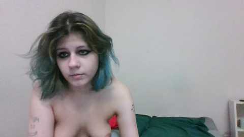 Media: A video of a topless young woman with shoulder-length, messy teal hair, wearing dark eye makeup, and visible tattoos. She sits on a bed with green and red sheets, in a dimly lit room with plain white walls.